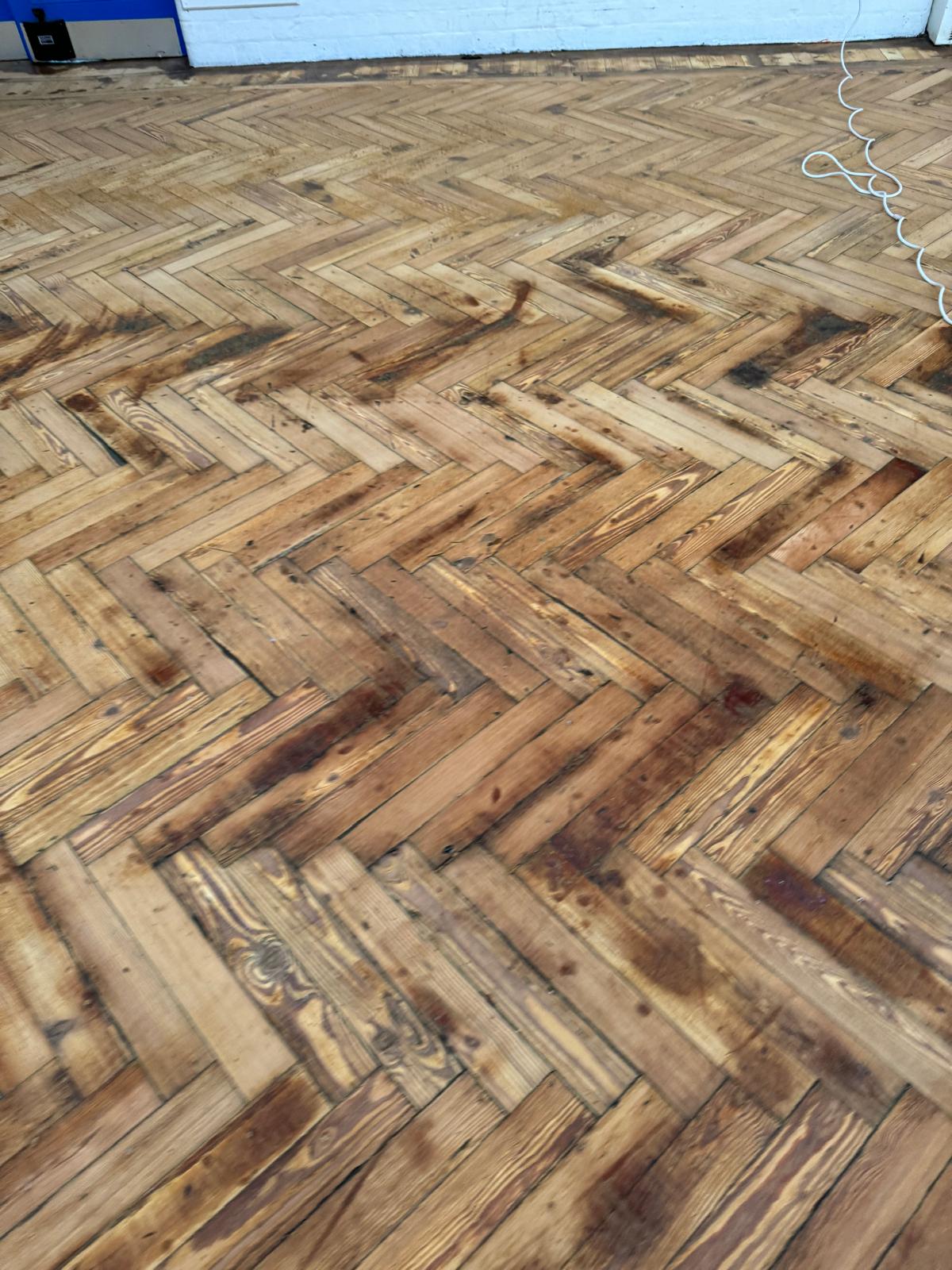 Part-sanded herringbone parquet floor during school hall sanding, showing fresh timber tones alongside darker, unsanded patches.