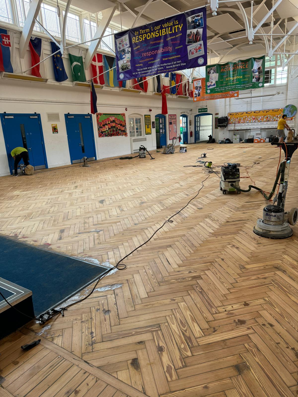 School hall sanding in progress: technicians manoeuvre dust-free drum and rotary sanders across a herringbone parquet floor beneath colourful international flags and “Responsibility” banners.